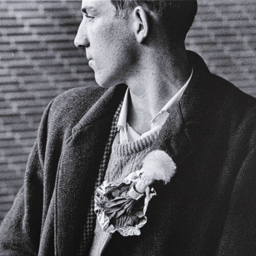 Black and white photograph of a man turned to look out a window. He wears a large brooch.